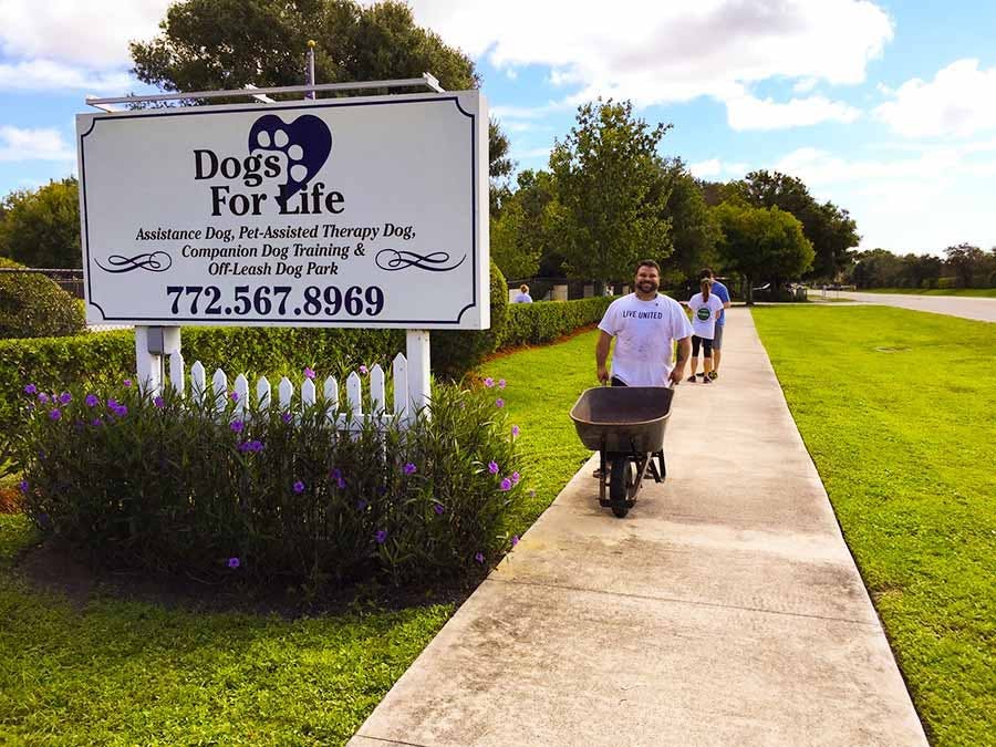 Volunteer pushing wheelbarrow on sidewalk by Dogs for Life sign 